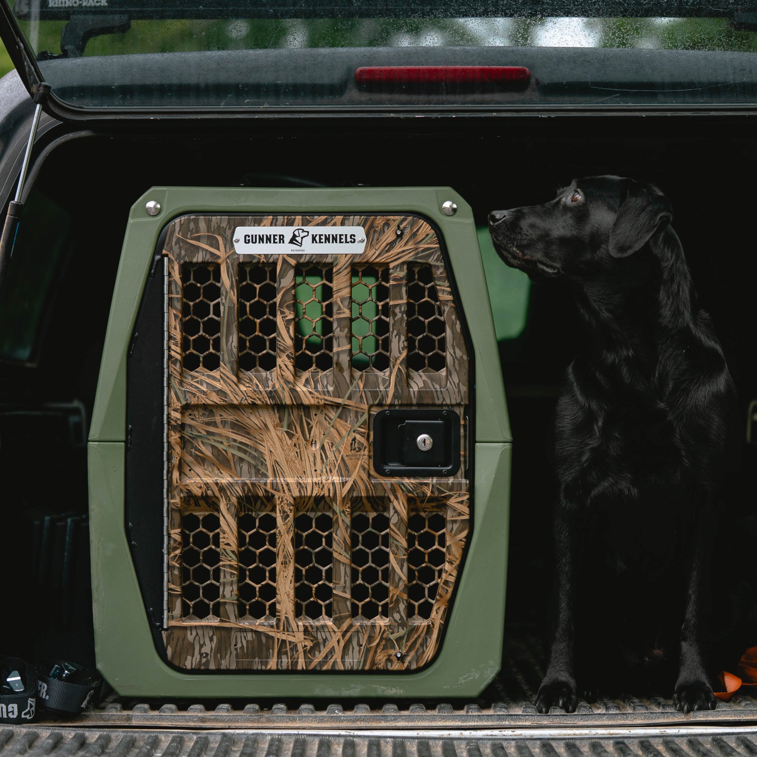 A black Labrador retriever sits in a vehicle next to the GUNNER G1™ KENNEL in Gunner Green/Mossy Oak Shadow Grass, a double-walled crate with a metal door. Sunlight highlights the camo-patterned kennel as the dog looks up.