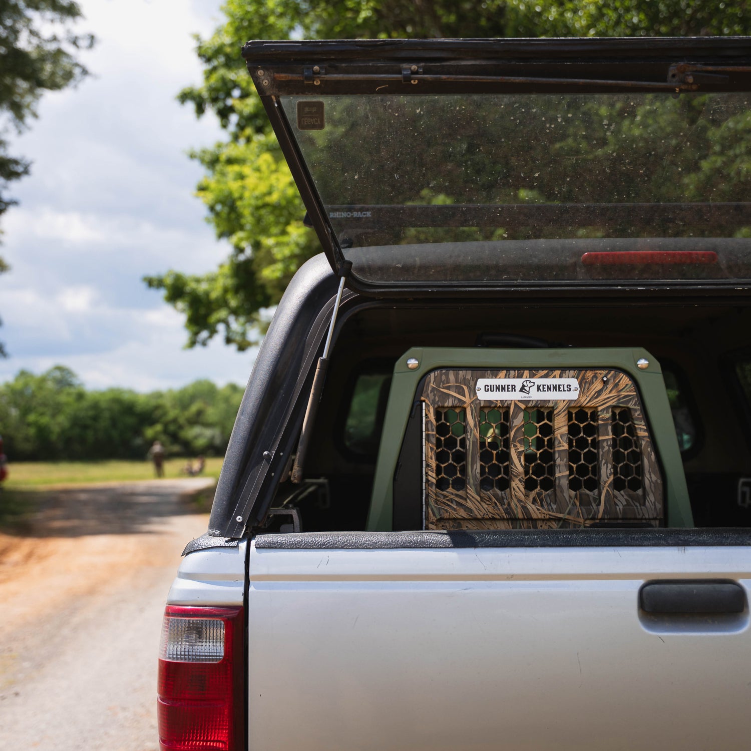 A pickup truck with its tailgate open reveals a GUNNER G1™ Kennel in Gunner Green/Mossy Oak Shadow Grass inside the truck bed, parked on a dirt road lined with trees under a partly cloudy sky.