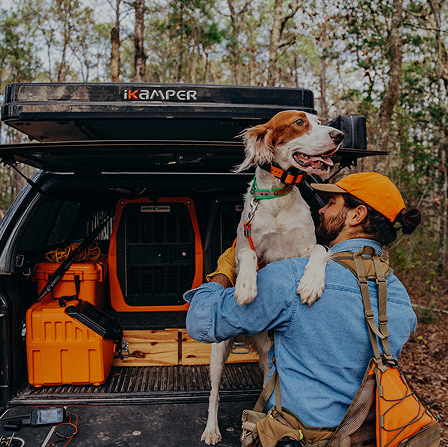 A man in an orange cap holds a white and brown hunting dog in front of a vehicle equipped with a G1™ KENNEL - BLAZE ORANGE in the back.
