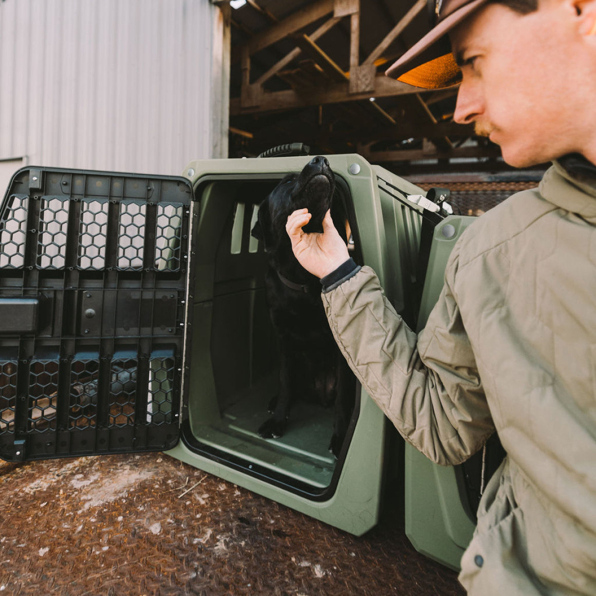 A man in a green jacket and brown cap gently pets a black dog sitting inside a GUNNER G1™ KENNEL in Gunner Green, one of the safest American-made crates, outdoors near a metal building.