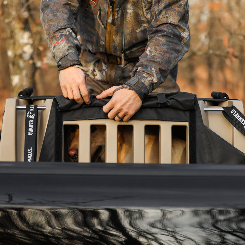 A person in camouflage clothing secures a black cover on a tan dog kennel, with a working dog visible inside. The scene is outdoors, with blurred trees in the background.