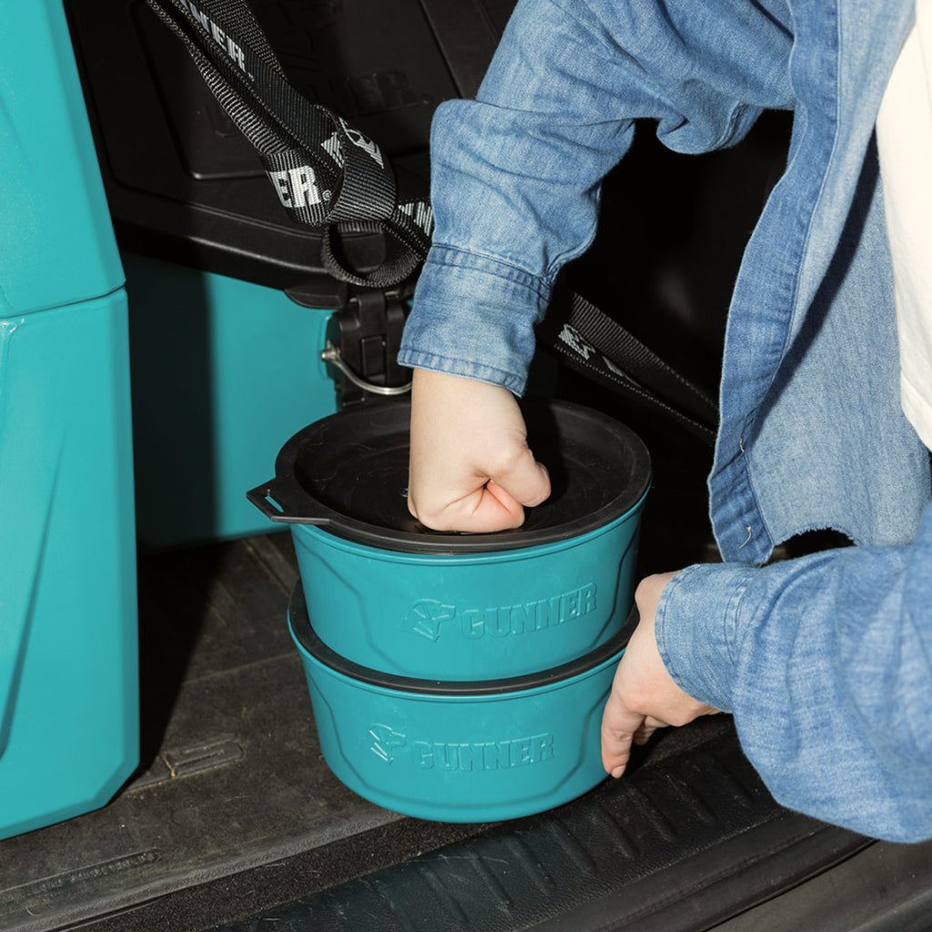 A person in a denim shirt lifts two teal GUNNER DOG BOWLs with black lids, stacked and labeled GUNNER, from a vehicle’s back seat—showcasing the Color Fetch 2025 collection.