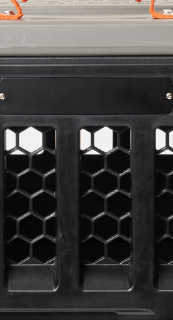 Close-up of a black plastic dog kennel or container with hexagonal mesh openings and a blank black nameplate, featuring orange straps at the top—perfect for safely transporting your hunting dog or working dog.