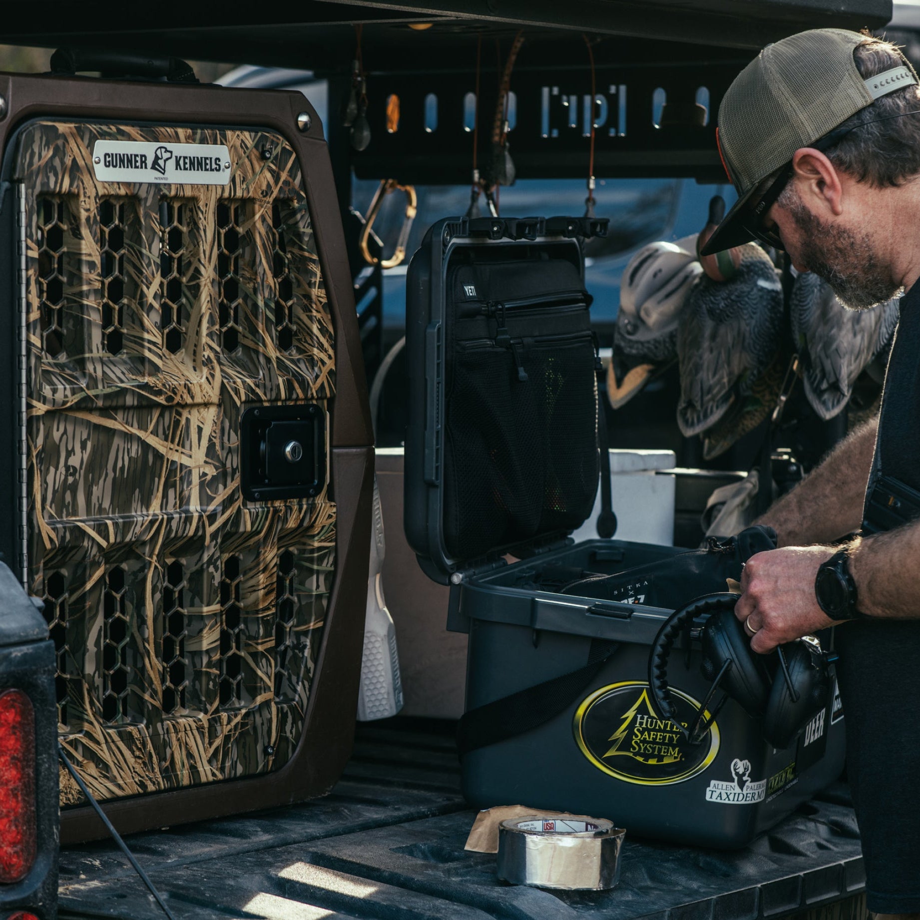 A man organizes gear in the back of a pickup beside a large GUNNER G1™ KENNEL - SUSQUEHANNA/MOSSY OAK SHADOW GRASS—perfect for duck and goose hunters—with outdoor equipment and a helmet visible in the truck bed.