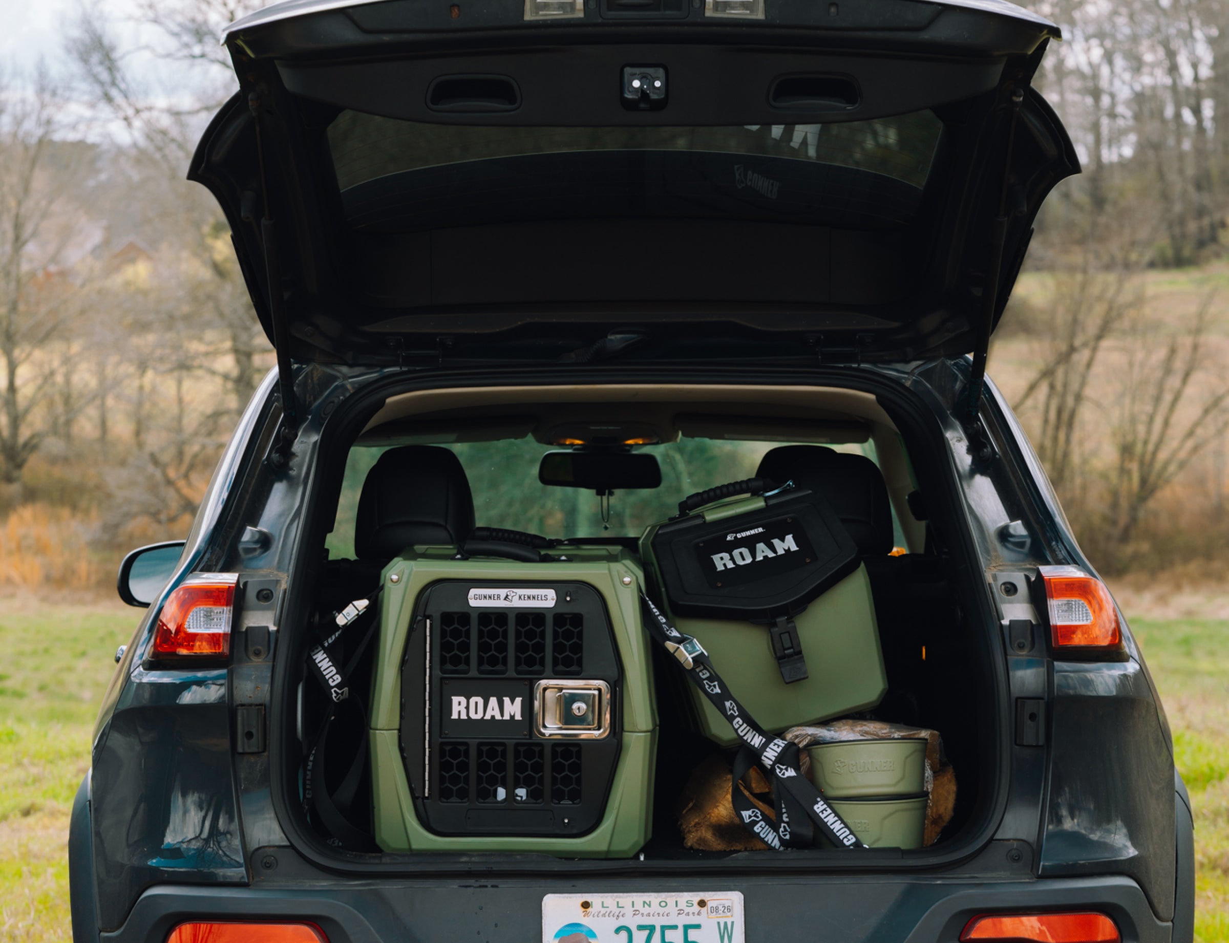 The open trunk of an SUV reveals GUNNER gear, including the G1™ KENNEL - GUNNER GREEN, arranged neatly. The car is parked outdoors with trees and grass in the background.