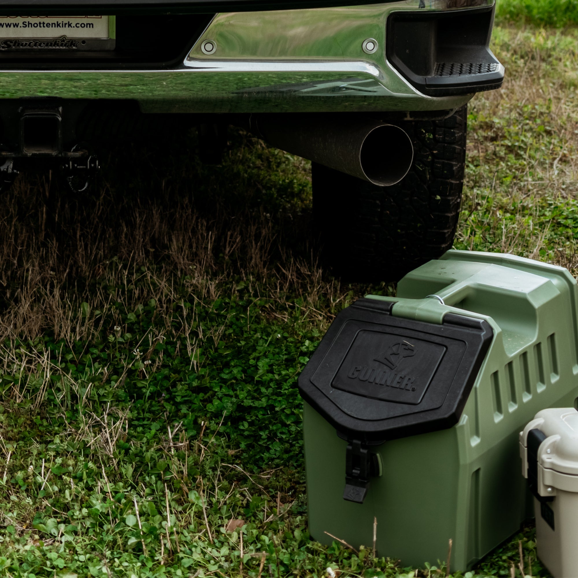 A GUNNER FOOD CRATE in GUNNER Green with a black lid sits on grass near a car’s bumper. Next to it is a partially visible, American-made dog food storage container.