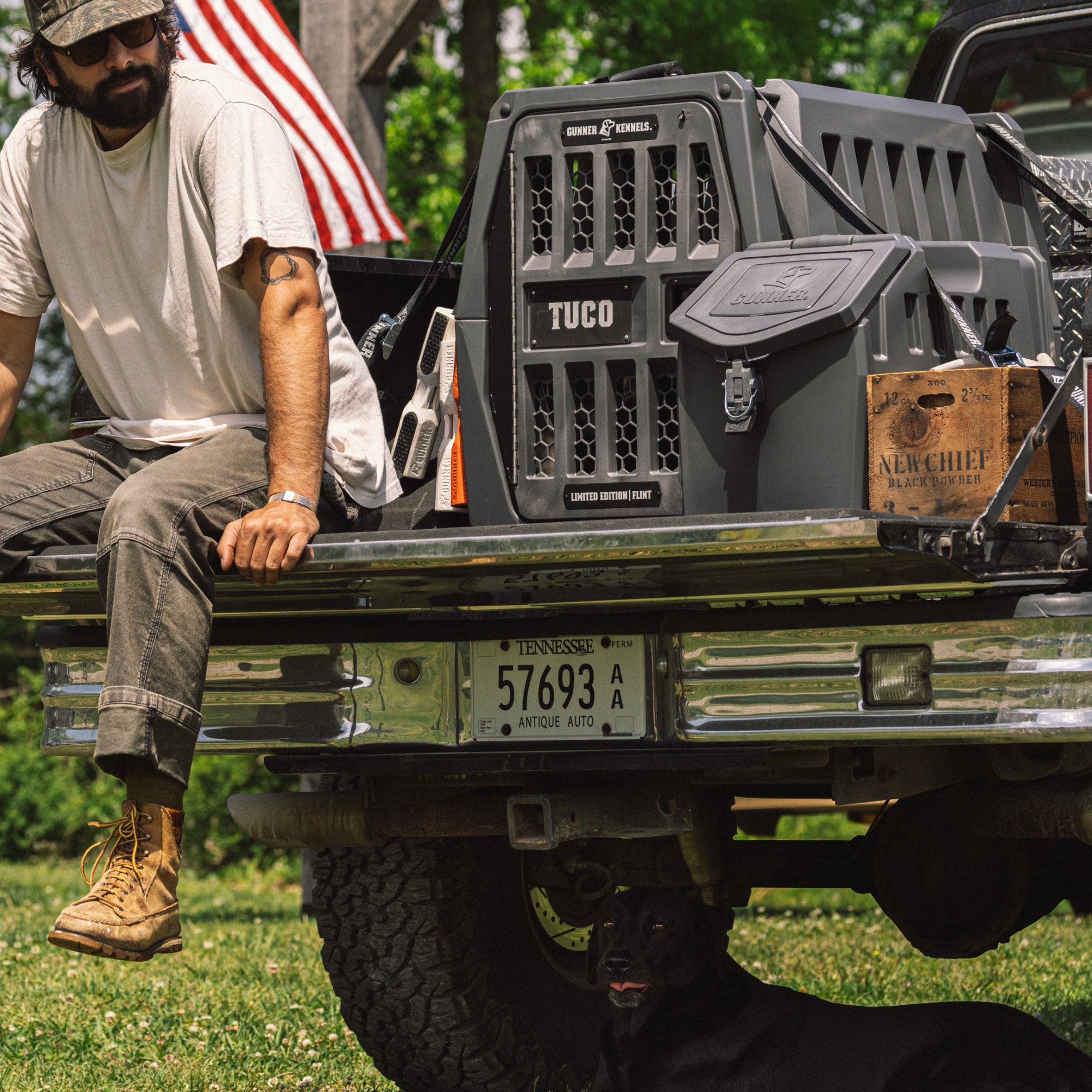 A man in work clothes and boots sits on a pickup tailgate with the GUNNER FOOD CRATE - FLINT by GUNNER. An American flag, license plate, and a black dog underneath highlight this Made in America scene.