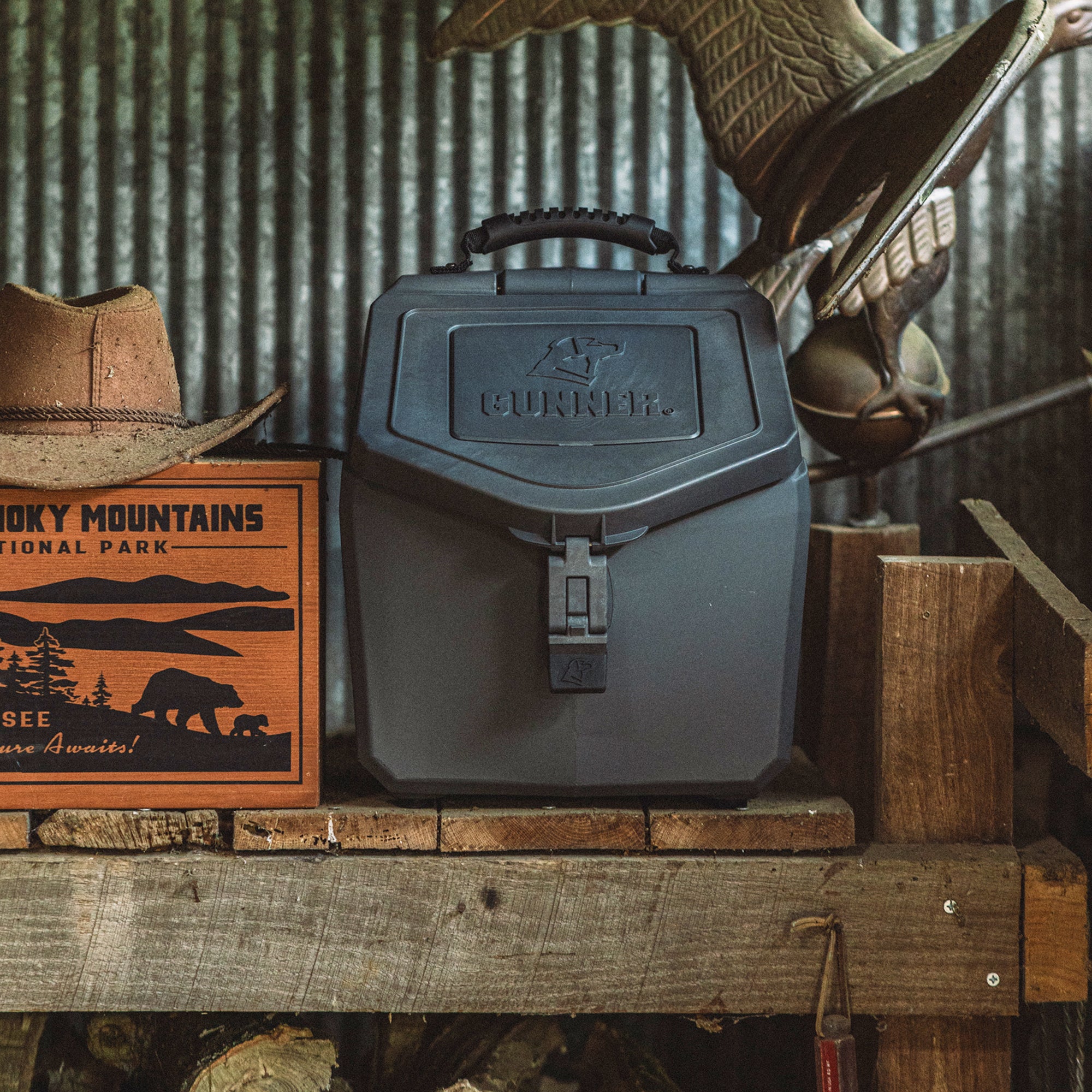 A GUNNER FOOD CRATE - FLINT, a black airtight dog food container by GUNNER, sits on a rustic wooden shelf beside a Smoky Mountains National Park sign and cowboy hat, with a metal eagle sculpture and corrugated metal wall behind.