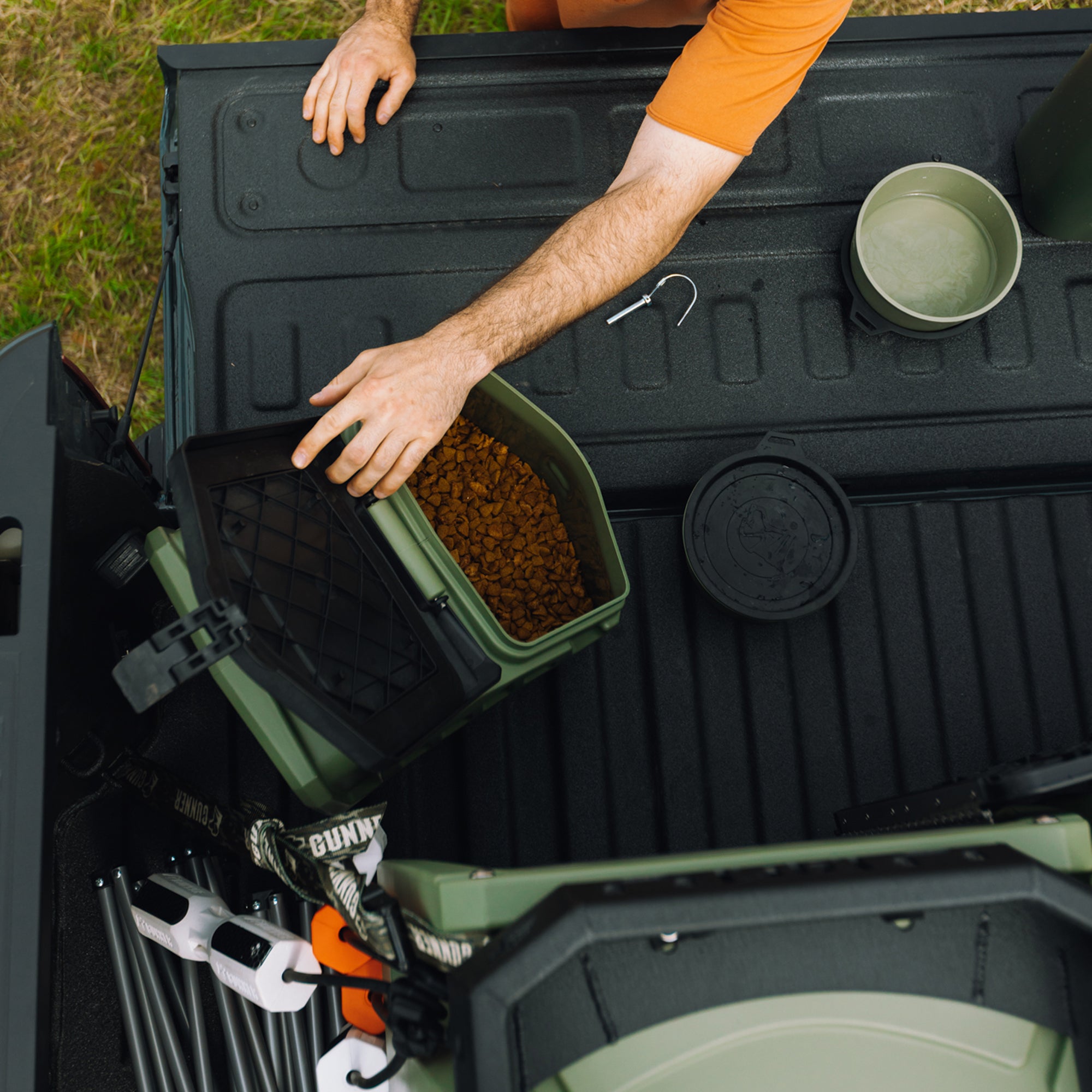 A person in an orange shirt reaches into a GUNNER FOOD CRATE - GUNNER Green filled with brown kibble on a truck bed. Nearby are a water bowl and tools on the textured surface. Made in America by GUNNER.