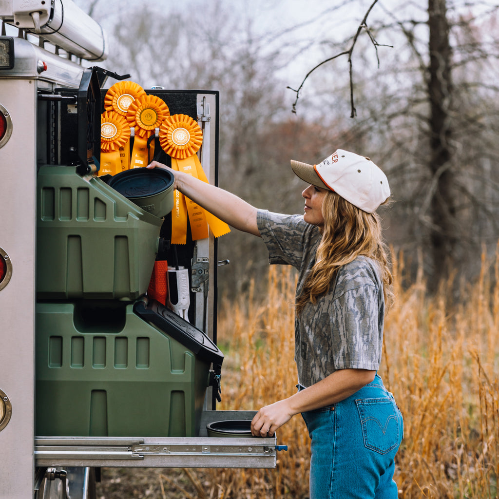A woman in a hat and denim jeans reaches into a truck storage compartment containing large orange award ribbons and the GUNNER FOOD CRATE - GUNNER Green by GUNNER, with tall grass and bare trees visible in the background.