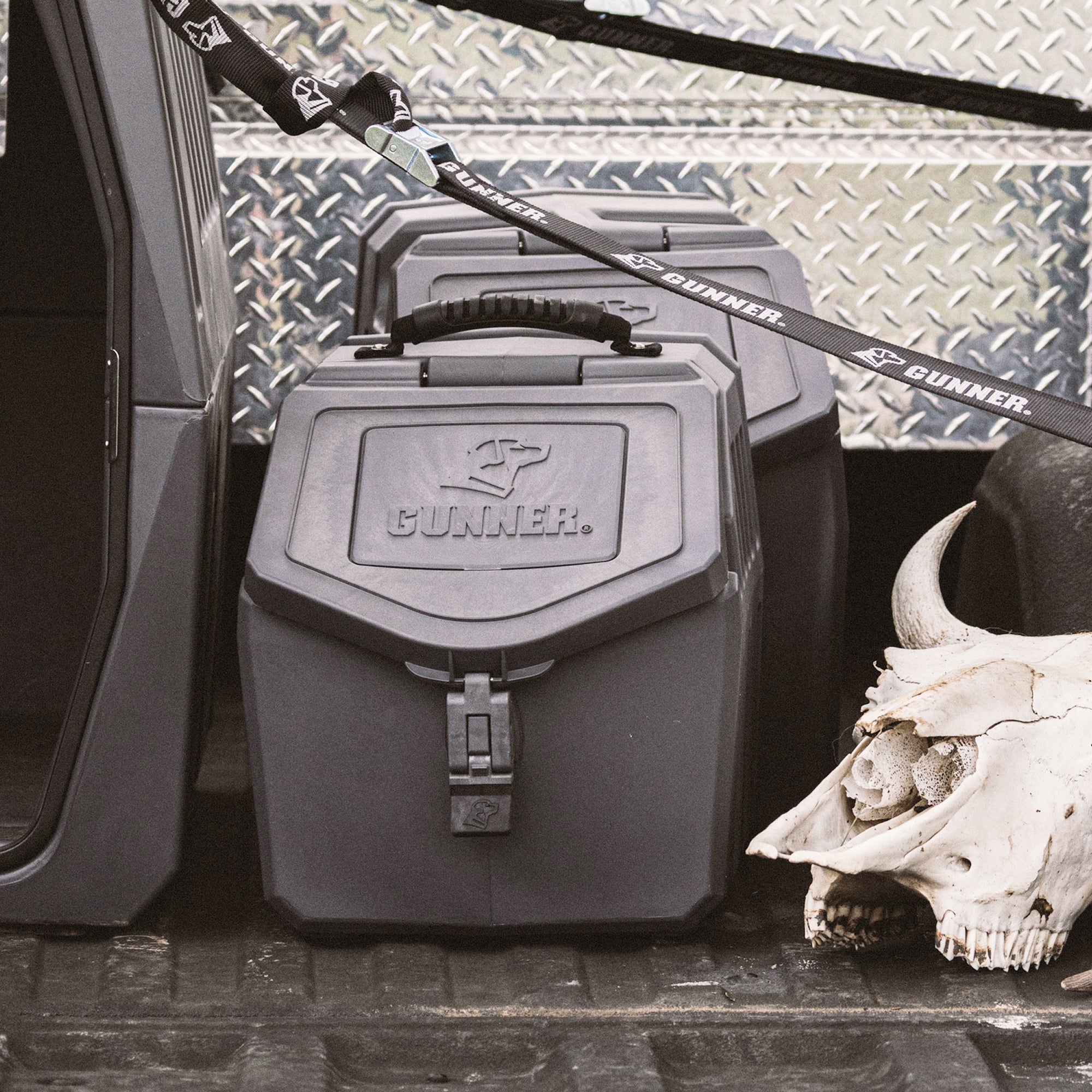 Two black GUNNER FOOD CRATE - FLINT airtight dog food containers with handles sit on a truck bed next to a cow skull, secured by a GUNNER-branded strap. Diamond plate metal is visible in the background. Made in America.