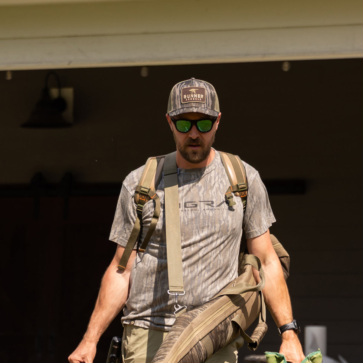 Man wearing a GUNNER BOTTOMLAND MALLARD TRUCKER hat, sunglasses, and a camouflage t-shirt, carrying a long gear bag over his shoulder.