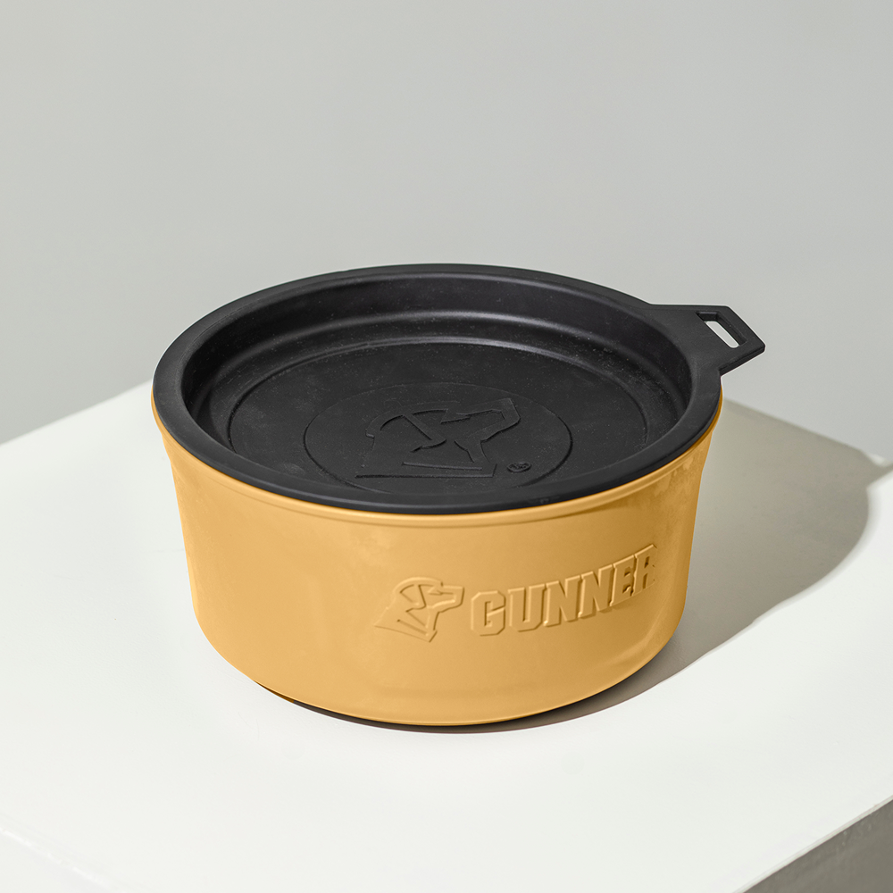 A tan and black round GUNNER DOG BOWL - Marigold with a lid, featuring the GUNNER logo on the side, is displayed on a white surface against a light gray background. It’s part of the Color Fetch 2025 collection.