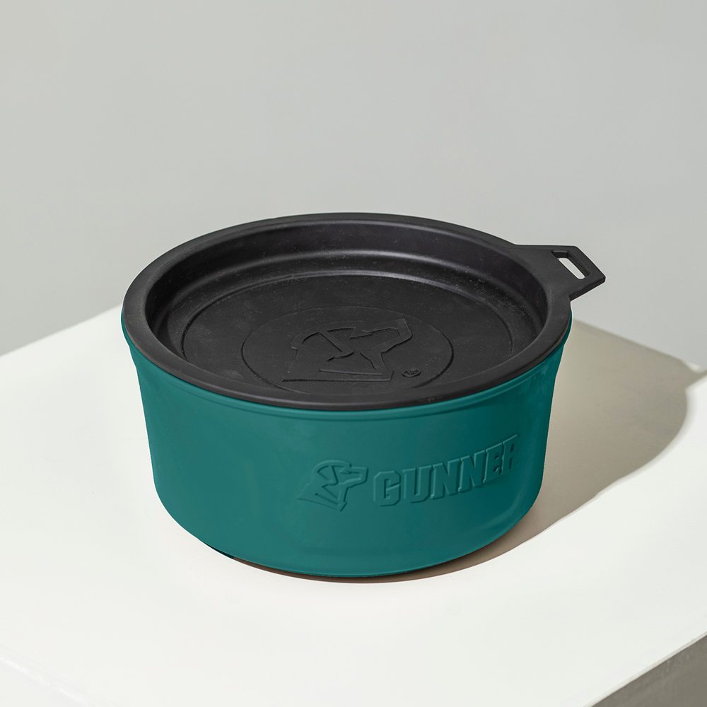 A teal and black round GUNNER DOG BOWL by GUNNER, featuring a handle on the lid and embossed logo, sits on a white surface—part of the stylish Color Fetch 2025 dog bowl collection.
