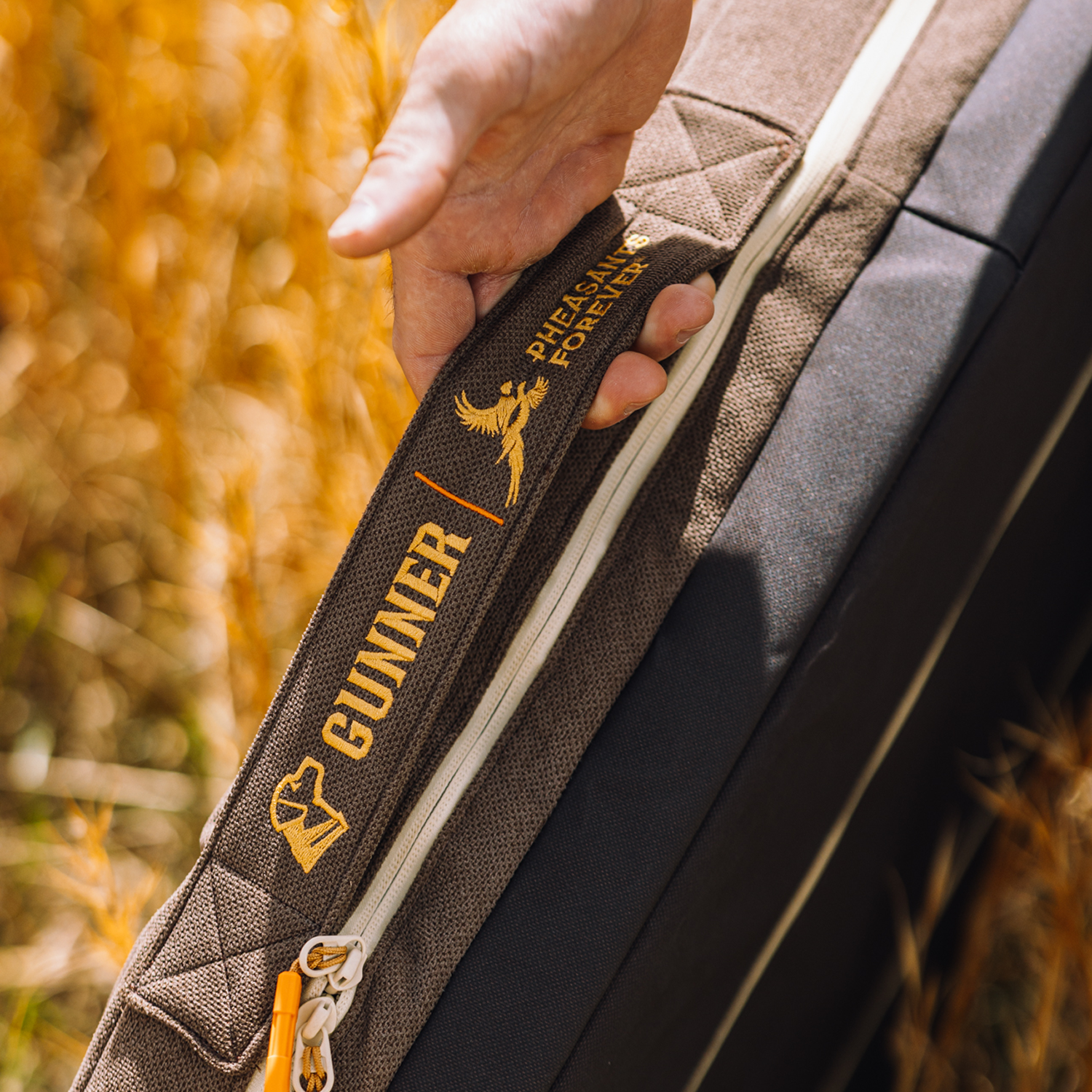 A hand grips a durable HOMESTEAD DOG BED in Special-Edition Pheasant Brown by GUNNER, with Pheasants Forever collaboration embroidered in gold. Golden reeds blur softly around the bed.