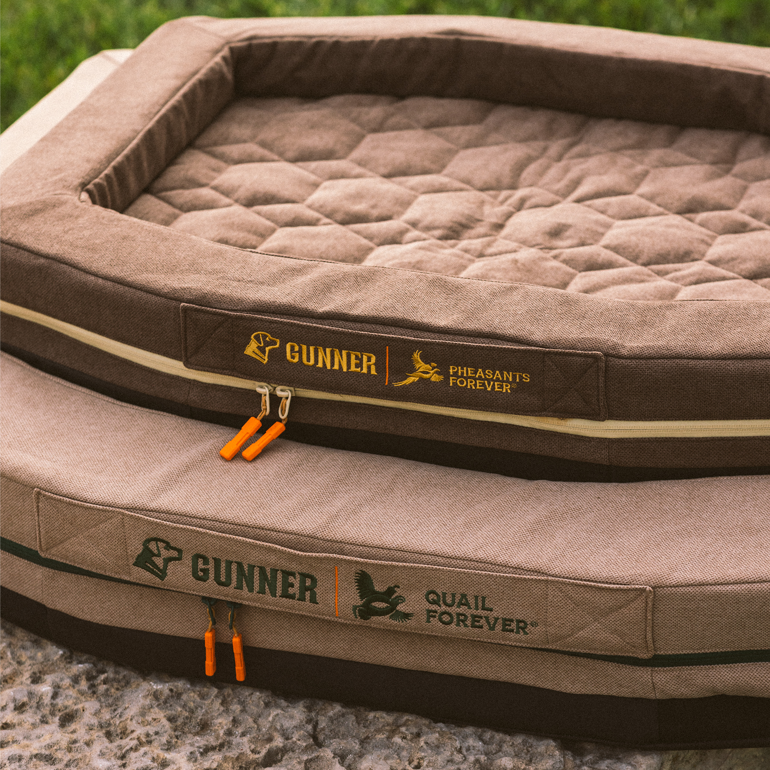Two brown, rectangular HOMESTEAD DOG BEDs by GUNNER sit outdoors. These Pheasant Brown beds have quilted tops, orange zipper pulls, and feature logos for GUNNER, Pheasants Forever, and Quail Forever.