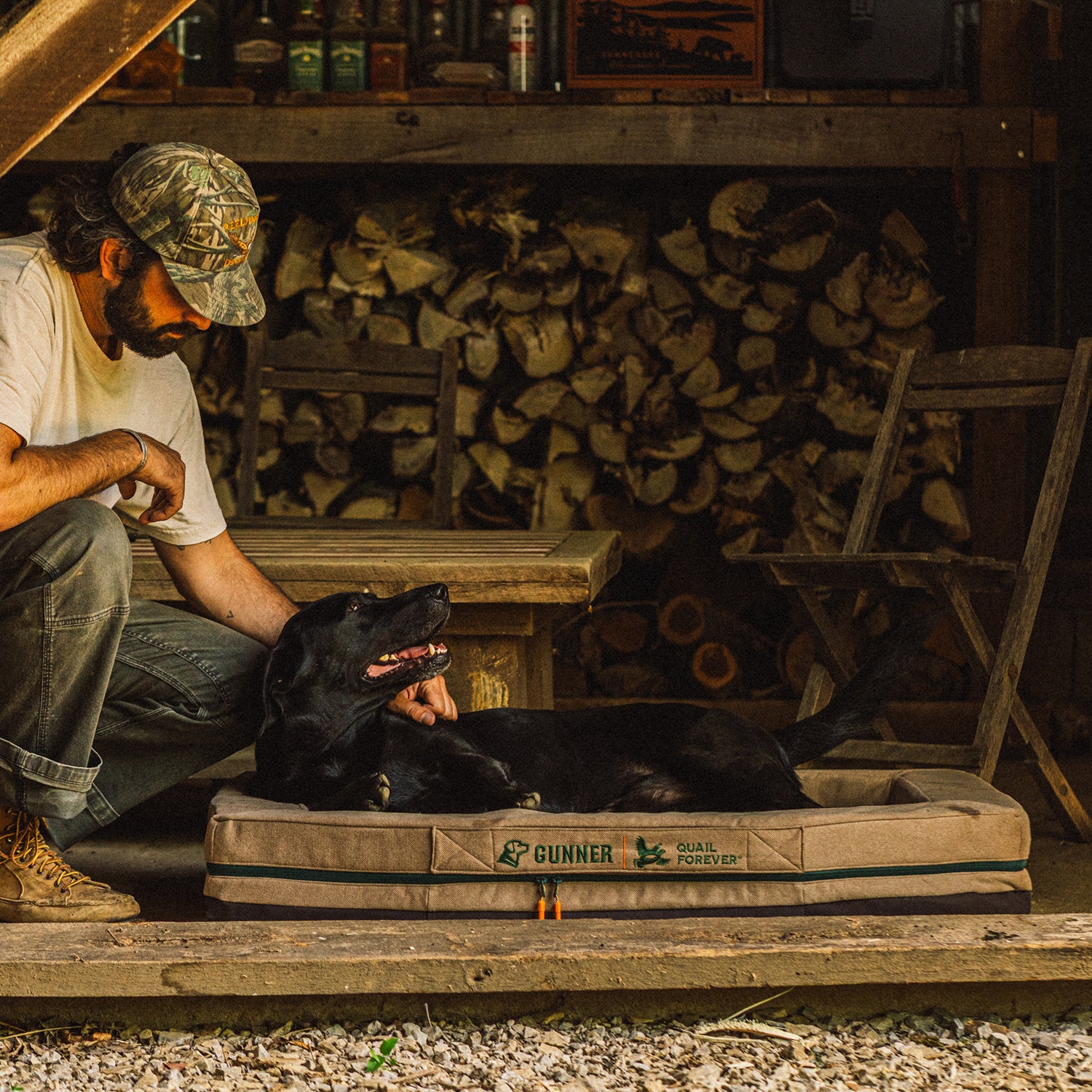 A man in a Special-Edition Quail Forever Tan camo cap pets a black dog lying in the GUNNER HOMESTEAD DOG BED Quail Tan on a wooden porch, with stacked firewood and rustic chairs in the background.