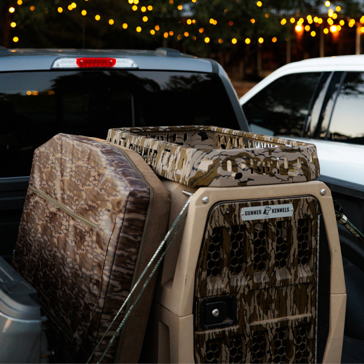 A Mossy Oak Bottomland GUNNER Kennel and matching cushion are securely strapped into the GUNNER Kennel Rack - Mossy Oak Bottomland in the bed of a pickup truck at dusk, with string lights glowing in the background. This setup maximizes truck bed real estate while keeping your gear organized.