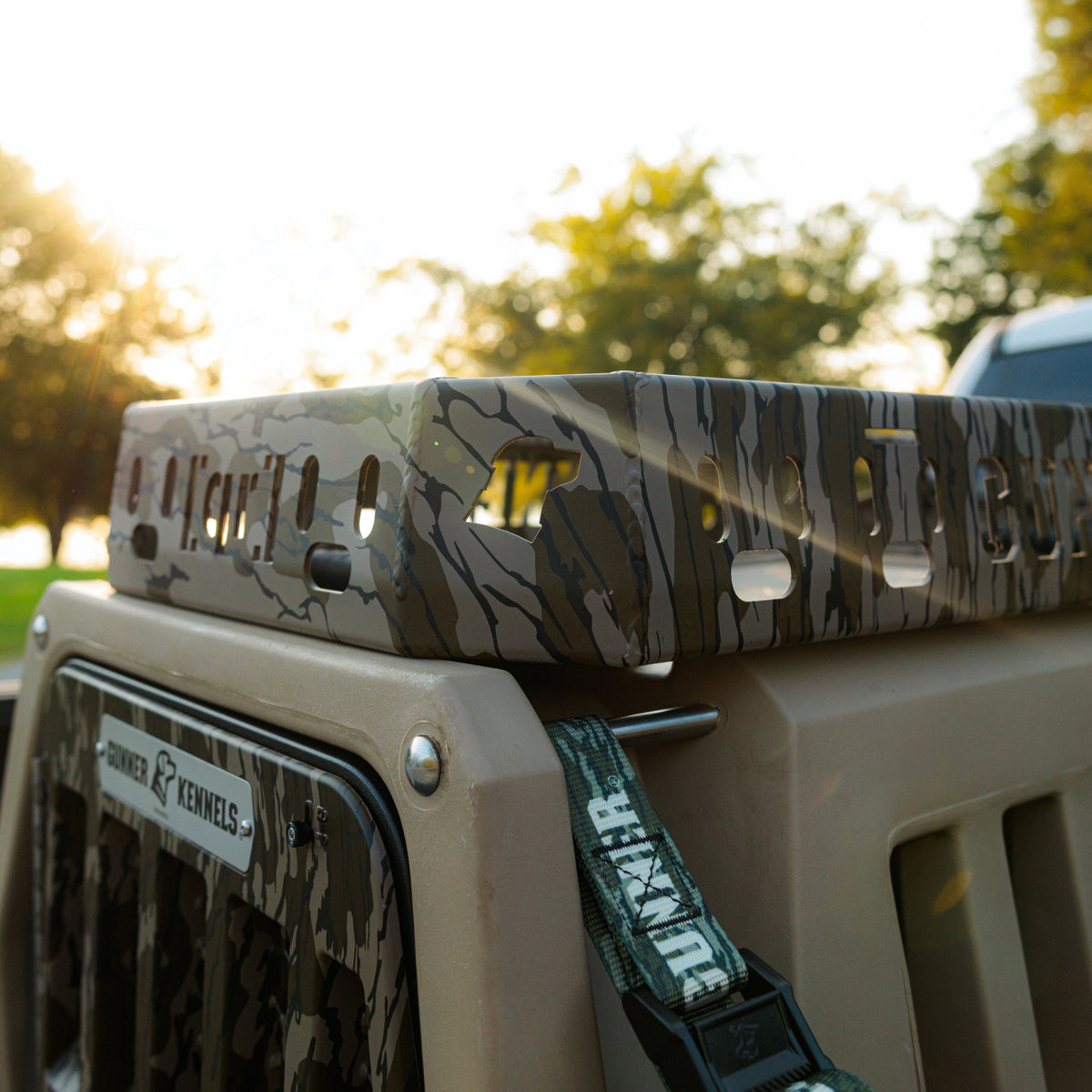 A GUNNER Kennel Rack in Mossy Oak Bottomland sits outdoors near a vehicle, sunlight filtering through trees in the background, with a green GUNNER-branded strap securing the setup. Designed to maximize your truck bed space, this stylish and organized solution is perfect for your next adventure.