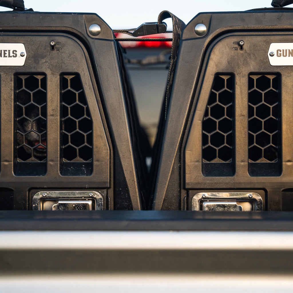 Two black G1™ KENNEL crates from GUNNER, each with hexagonal ventilation holes, are secured side by side in a vehicle. A dog is seen in the left crate. Both tough crates display the GUNNER brand name.