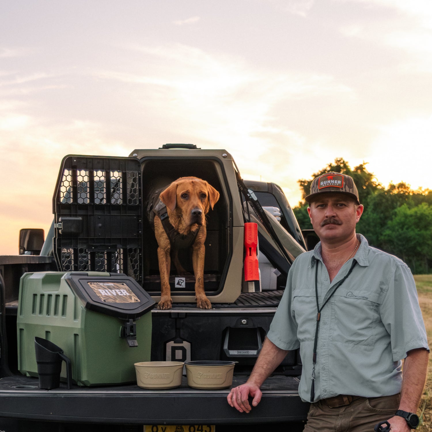 A man in outdoor gear stands next to a pickup truck at sunset, with his brown dog resting on a GUNNER LONG HAUL BED inside a crate in the truck bed. Camping gear and dog bowls sit nearby, with trees and sky in the background.