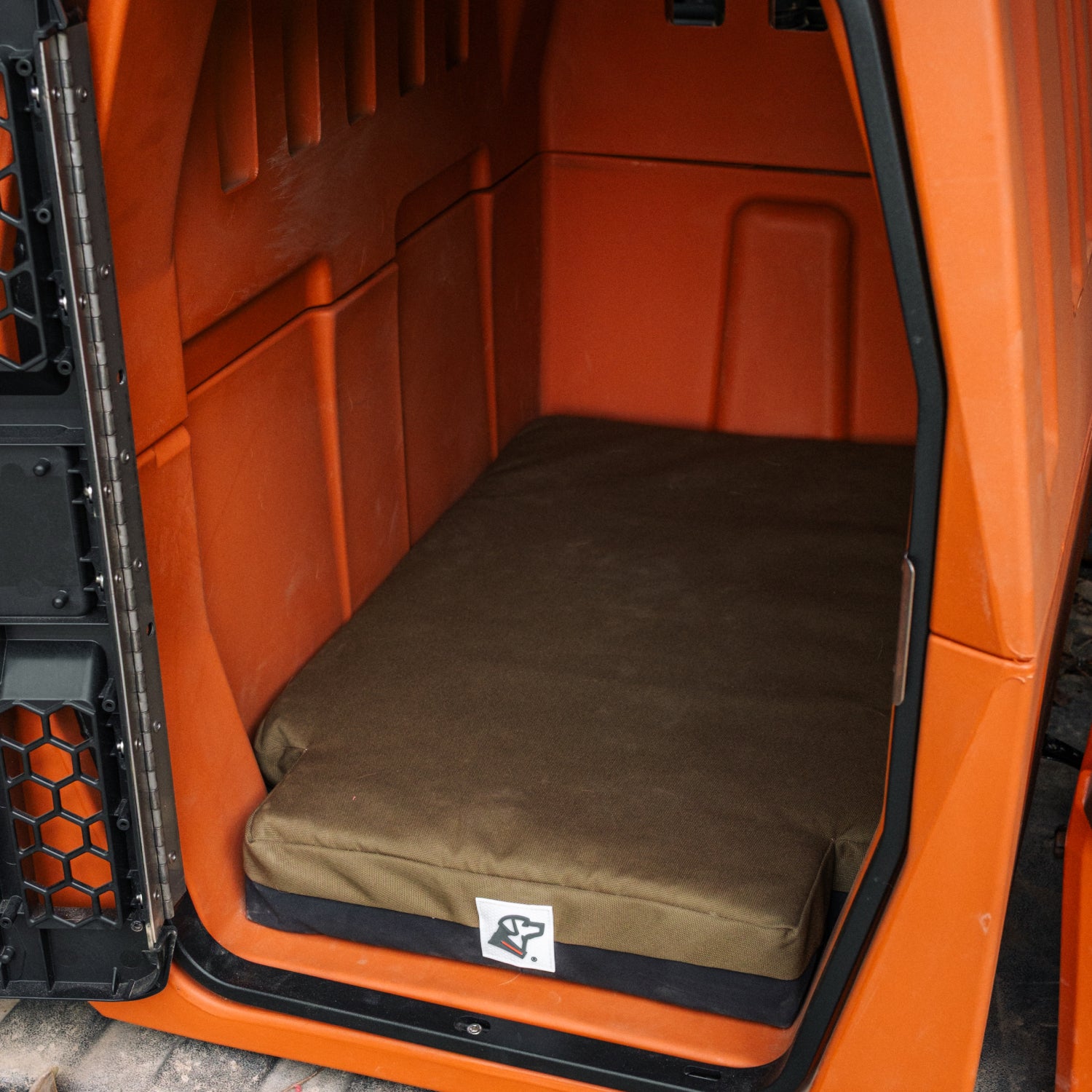 A GUNNER orange hard-sided kennel featuring the LONG HAUL BED, a brown, durable dog bed. The open door reveals a spacious interior and a hexagonal-patterned metal gate on the left.