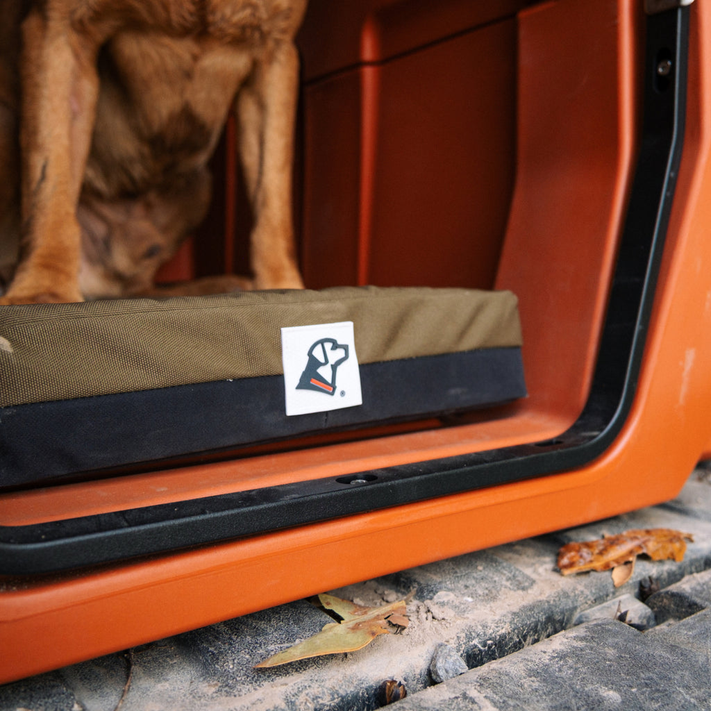 A brown dog stands on the GUNNER LONG HAUL BED, an olive and black waterproof kennel bed with a white heart and paw logo, inside an orange vehicle. The bed keeps fur dry while fallen leaves and dirt are visible outside.