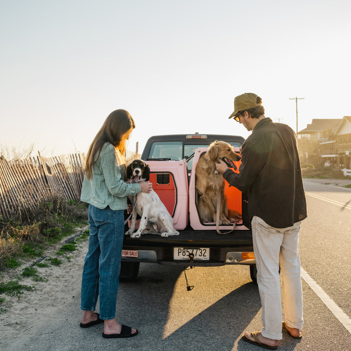 A woman and a man stand at the open tailgate of a parked pickup truck, each with a dog and some outdoor gear. The sun is setting, and they’re by a fence on a quiet road near houses.