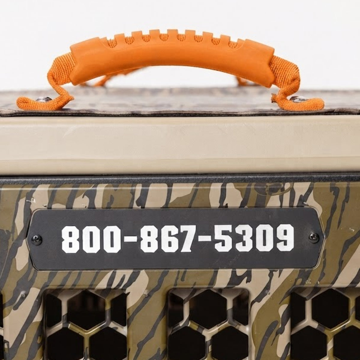 A camouflage-patterned dog kennel with an orange handle and a black sign displaying the phone number 800-867-5309, perfect for keeping your hunting dog secure while you’re on the go. This sturdy crate doubles as essential outdoor gear for adventurous pet owners.