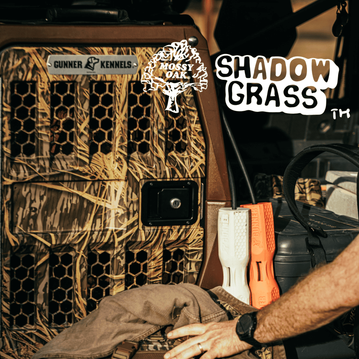A person’s arm rests near a camouflage-patterned dog kennel with the logo “Mossy Oak Shadow Grass,” orange and white dog training dummies, and essential outdoor gear attached to the side of a vehicle.