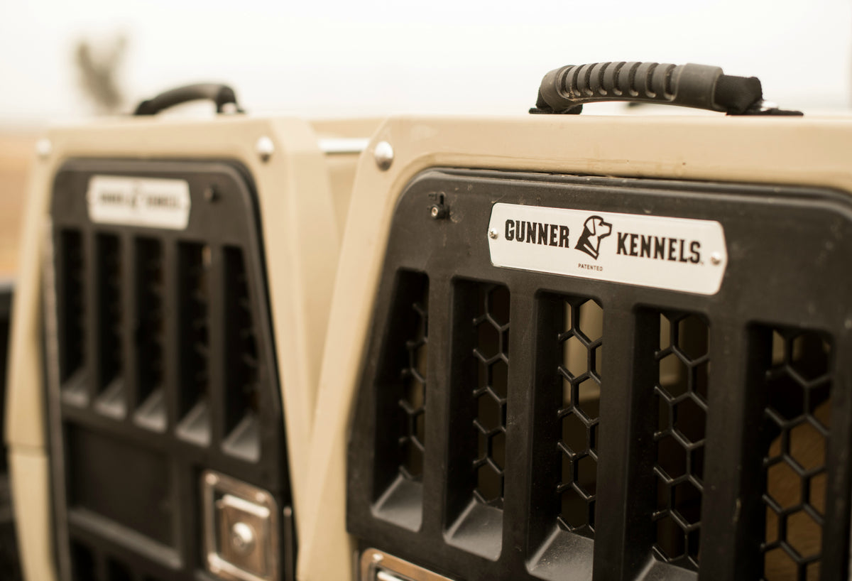 Close-up of two beige dog crates with hexagonal ventilation holes and handles on top, labeled Gunner Kennels. Ideal for transporting your working or hunting dog. The background is blurred.