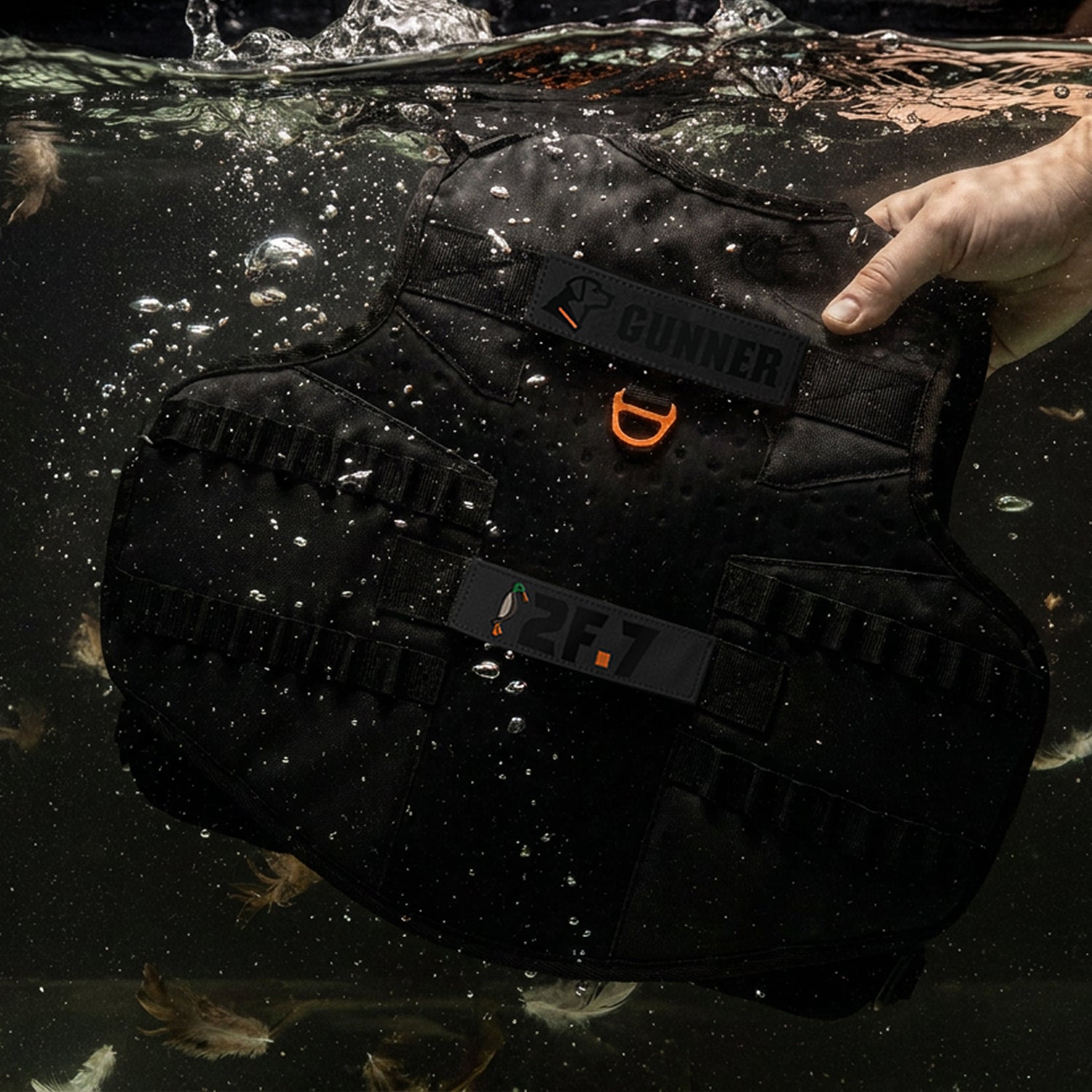 A hand holds the GUNNER x 2F.7 Waterfowl Dog Vest by GUNNER, black in color and partially submerged in water, with bubbles rising and a few feathers floating nearby.