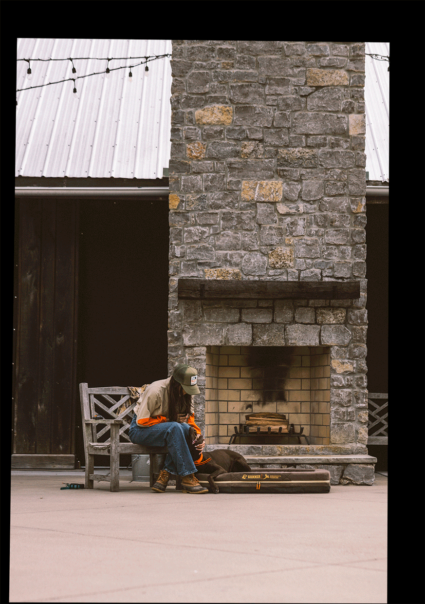 A woman sitting on a hair rubbing a dog while the dog lays on a dog bed in front of an outdoor fireplace.