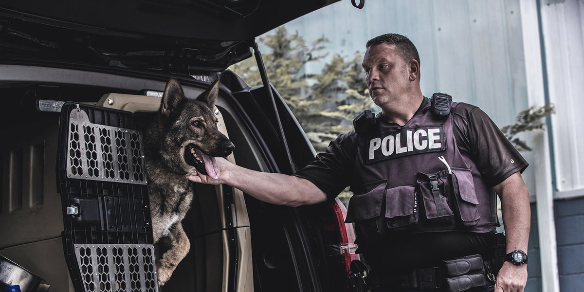 A police officer in a vest labeled POLICE stands beside an open vehicle, gently holding the mouth of a German Shepherd police dog that is partly inside a dog crate in the back of the car.